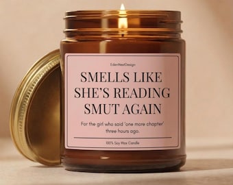 May include: A brown glass jar candle with a lit wick and a gold lid placed to the side. The label is light pink with black text that reads: EdenNestDesign, SMELLS LIKE SHE'S READING SMUT AGAIN, For the girl who said 'one more chapter' three hours ago, 100% Soy Wax Candle.
