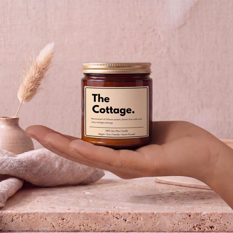 May include: A brown glass jar candle with a gold lid and a cream-colored label. The label features the text 'The Cottage.' followed by 'Reminiscent of Ottawa sunsets, distant loon calls and cozy cottage evenings.' and '100% Soy Wax Candle. Vegan &bull; Eco-Friendly &bull; Hand-Poured'.