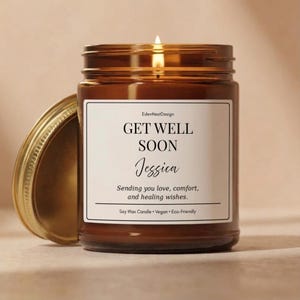 May include: A brown glass jar candle with a lit wick and a gold lid placed to the side. The white label features the text: EdenNestDesign, GET WELL SOON, Jessica, Sending you love, comfort, and healing wishes. Soy Wax Candle, Vegan, Eco-Friendly.