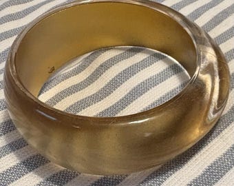 May include: A wide, gold-coloured bangle bracelet. The bracelet has a smooth, polished surface and a rounded shape. The interior of the bracelet is a lighter gold colour. The background is a blue and white striped fabric.
