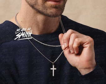 May include: A man wearing a dark blue sweater with a silver chain necklace and a silver cross pendant. The necklace has the text "We are all Smith" on it.