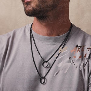 May include: A man wearing a gray t-shirt with the text "We are all Smith" printed on it. He is wearing two black necklaces with circular pendants.
