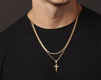 May include: A gold-colored necklace set featuring two chains. The top chain is a thinner rope style, and the bottom chain has a cross pendant. The person in the image is wearing a black t-shirt.