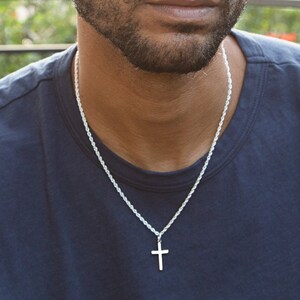 May include: A man with short, dark hair and a beard is wearing a dark blue t-shirt and a silver chain necklace with a small silver cross pendant.