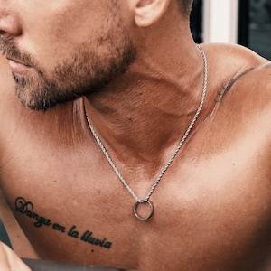 May include: Silver-toned necklace with a circular pendant. The necklace is worn on a person's bare chest, with a tattoo that reads "Danza en la lluvia". The person is leaning on a black bar.