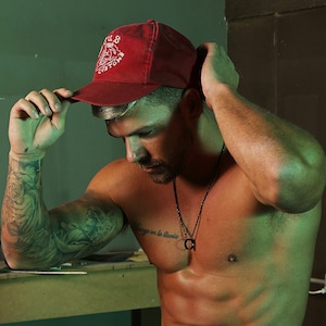 May include: A man wearing a red baseball cap with the text "VILLOS CUSTOM" in white, a black necklace with a circular pendant, and ripped black jeans. He is sitting on a workbench with a green background.