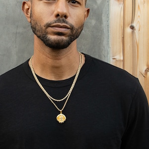 May include: A man wearing a black t-shirt with a gold chain necklace and a gold pendant.