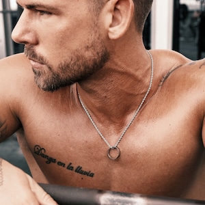 May include: A shirtless man with a silver chain necklace and a tattoo on his chest that reads "Danza en la lluvia".