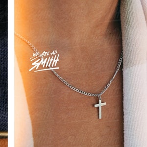 May include: A silver chain necklace with a small silver cross pendant. The necklace is worn by a person with a white towel draped over their shoulder. The text "We Are All Smith" is written in white on the image.