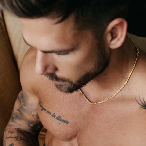 May include: A man with a beard and tattoos is wearing a gold chain necklace. The chain is a rope style and is visible against his bare chest. The man has a tattoo on his chest that says "Danza en la lluvia".