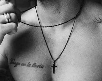 May include: A black chain necklace with a cross pendant. The necklace is worn by a person with a tattoo on their chest that reads "Danza en la lluvia".