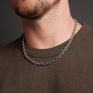 May include: A silver-colored chain necklace with a rounded link design. The necklace is worn over a dark olive-green t-shirt. The image is a close-up of the necklace and upper chest area.