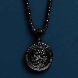 May include: Black chain necklace with a round pendant featuring a silver image of Saint Christopher and the text "Saint Christopher Protect Us".