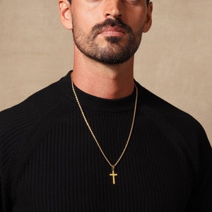 May include: A gold-colored cross pendant hangs from a gold chain. The cross is a simple, classic design. The chain is worn over a black ribbed sweater.