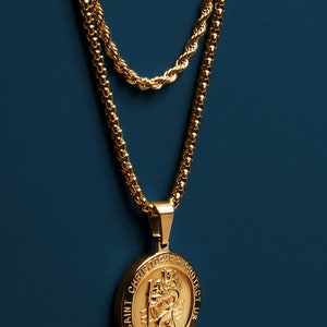 May include: A gold chain necklace with a gold pendant featuring the image of Saint Christopher and the text "Saint Christopher Protect Us".