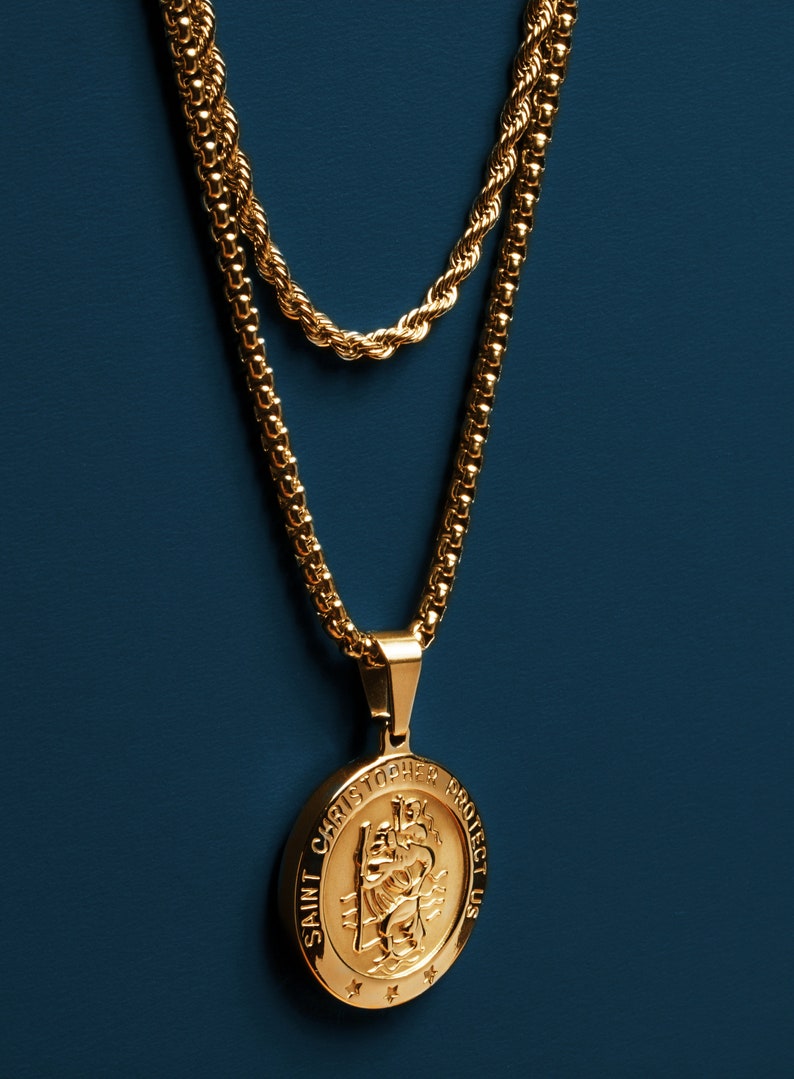May include: A gold chain necklace with a gold pendant featuring the image of Saint Christopher and the text "Saint Christopher Protect Us".