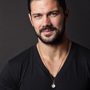May include: A man with short, dark brown hair and a beard is wearing a black v-neck t-shirt and a gold chain necklace with a compass pendant.