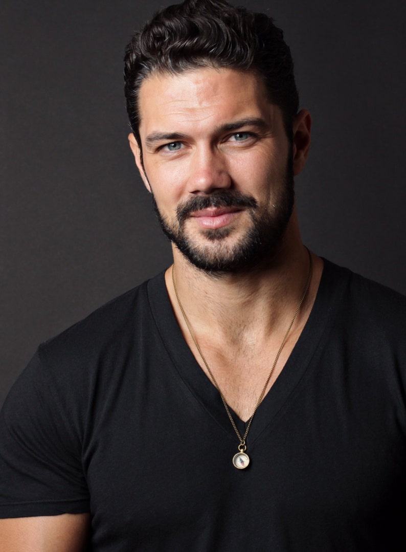 May include: A man with short, dark brown hair and a beard is wearing a black v-neck t-shirt and a gold chain necklace with a compass pendant.