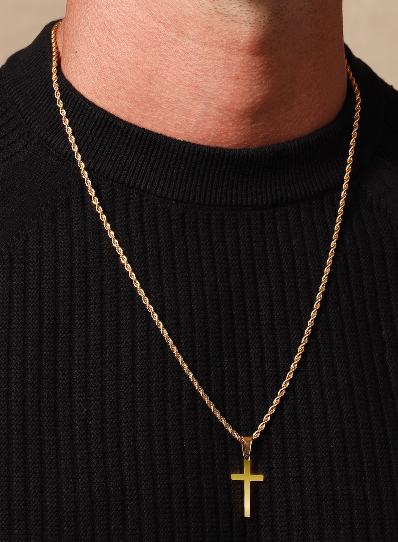 May include: A gold-colored cross pendant necklace. The cross hangs from a matching gold rope chain. The necklace is displayed against a black background.