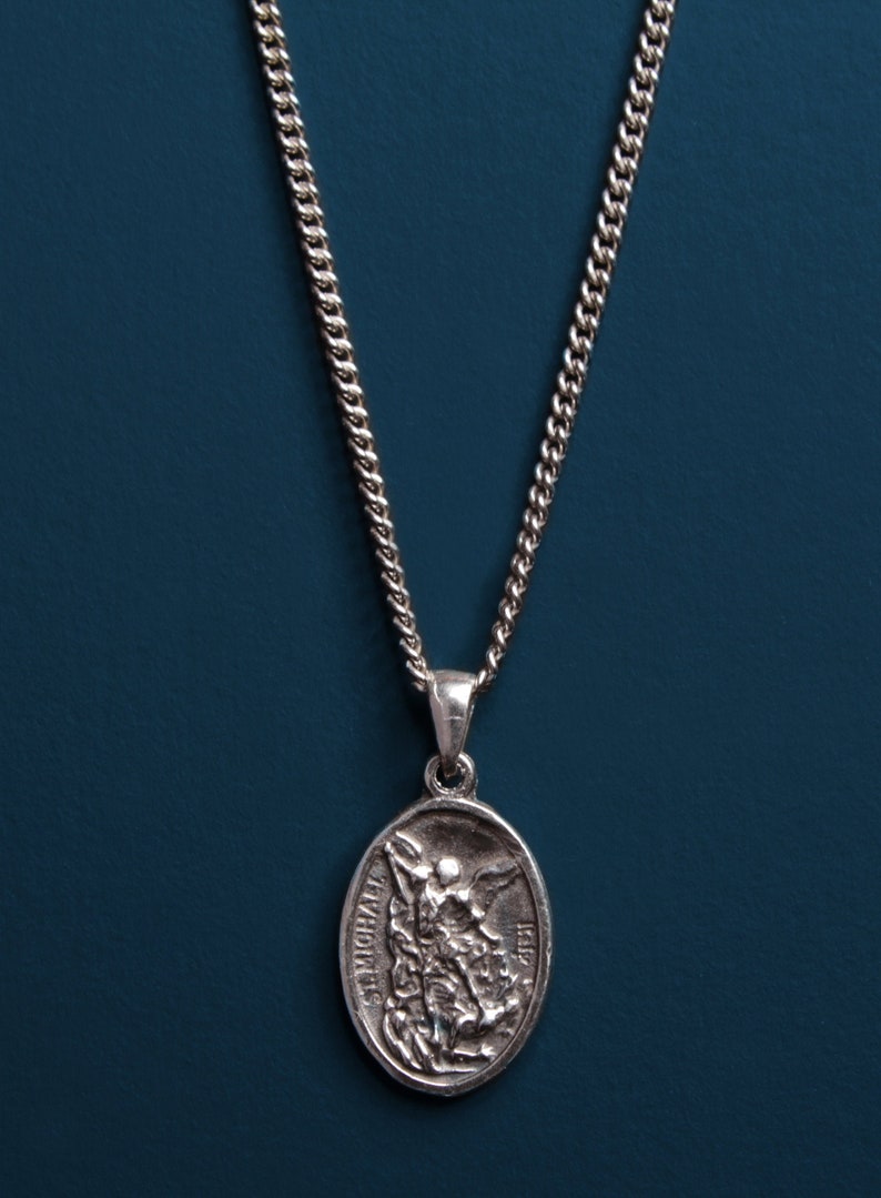 Pu&ograve; includere: Una collana con catena d'argento con un ciondolo ovale in argento raffigurante un angelo con le ali. Il ciondolo ha le parole "St. Michael" e "1518" incise.