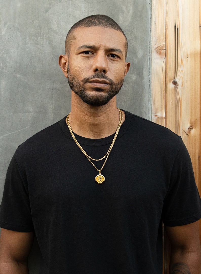 May include: A man wearing a black t-shirt with a gold chain necklace and a gold pendant.