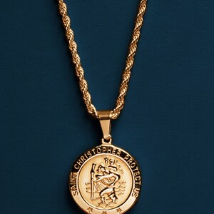 May include: A gold chain necklace with a round pendant featuring the image of Saint Christopher and the text "Saint Christopher Protect Us".