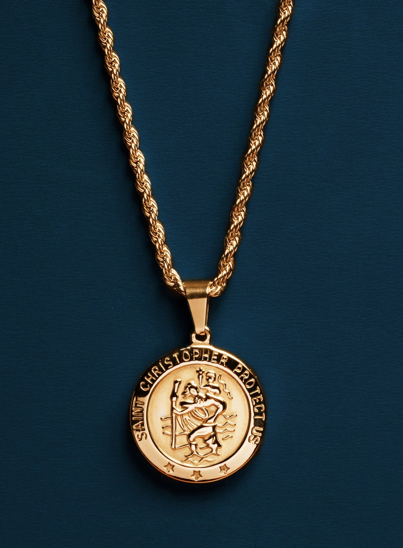 May include: A gold chain necklace with a round pendant featuring the image of Saint Christopher and the text "Saint Christopher Protect Us".