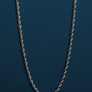 May include: Silver-toned rope chain necklace against a dark teal background. The chain is made of tightly twisted links, creating a rope-like texture. The necklace hangs vertically, showcasing its design and craftsmanship.