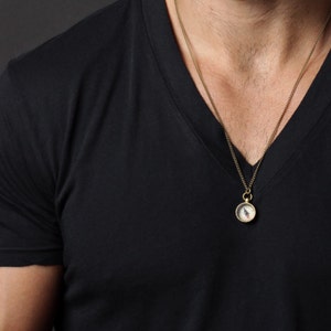 May include: A gold chain necklace with a small compass pendant. The compass is round and has a white face with black markings. The necklace is worn by a person wearing a black v-neck shirt.