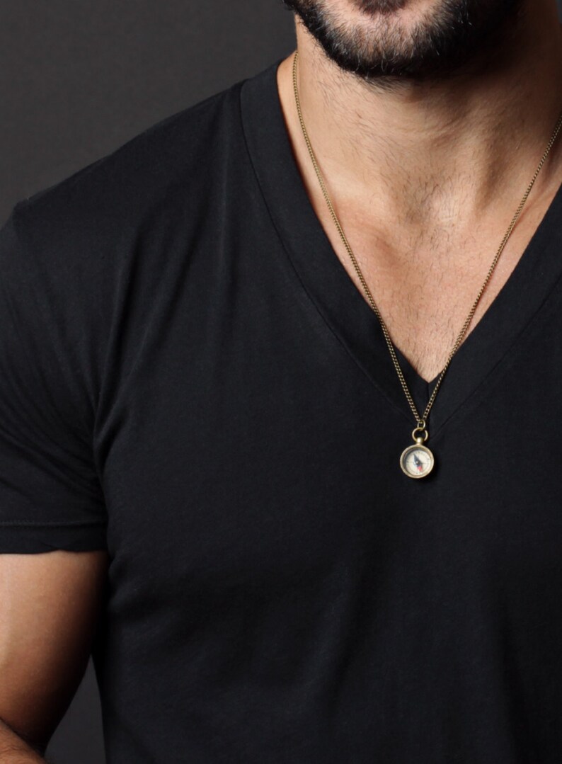 May include: A gold chain necklace with a small compass pendant. The compass is round and has a white face with black markings. The necklace is worn by a person wearing a black v-neck shirt.