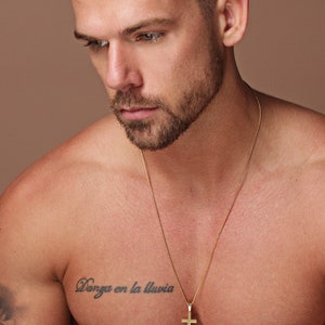 May include: A man wearing a gold chain necklace with a large gold cross pendant. The necklace is 26 inches long. The man has a tattoo on his chest that says "Danza en la lluvia".