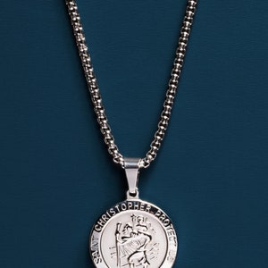 May include: A silver chain necklace with a round pendant featuring the image of Saint Christopher and the text "Saint Christopher Protect Us".
