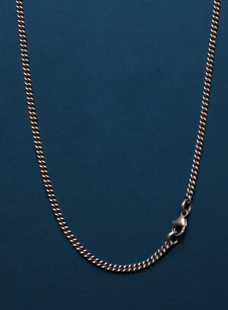 Pu&ograve; includere: Una collana a catena d'argento con chiusura a moschettone. La catena &egrave; su uno sfondo blu.