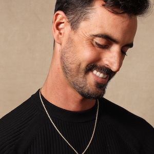 May include: A man wearing a black sweater with a gold rope chain necklace and a small gold cross pendant.