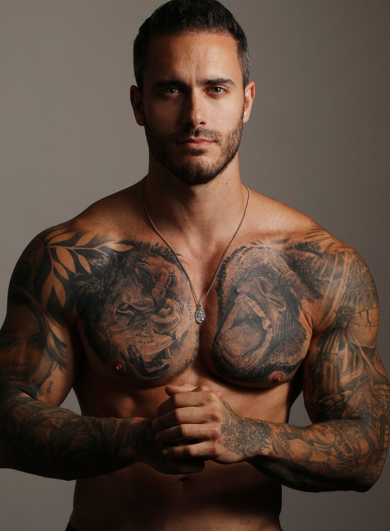 Pu&ograve; includere: Un uomo con la barba corta e capelli corti e scuri indossa una collana a catena d'argento con un ciondolo. Ha tatuaggi sul petto, sulle braccia e sulle spalle. I tatuaggi includono un leone, un gorilla e altri disegni.
