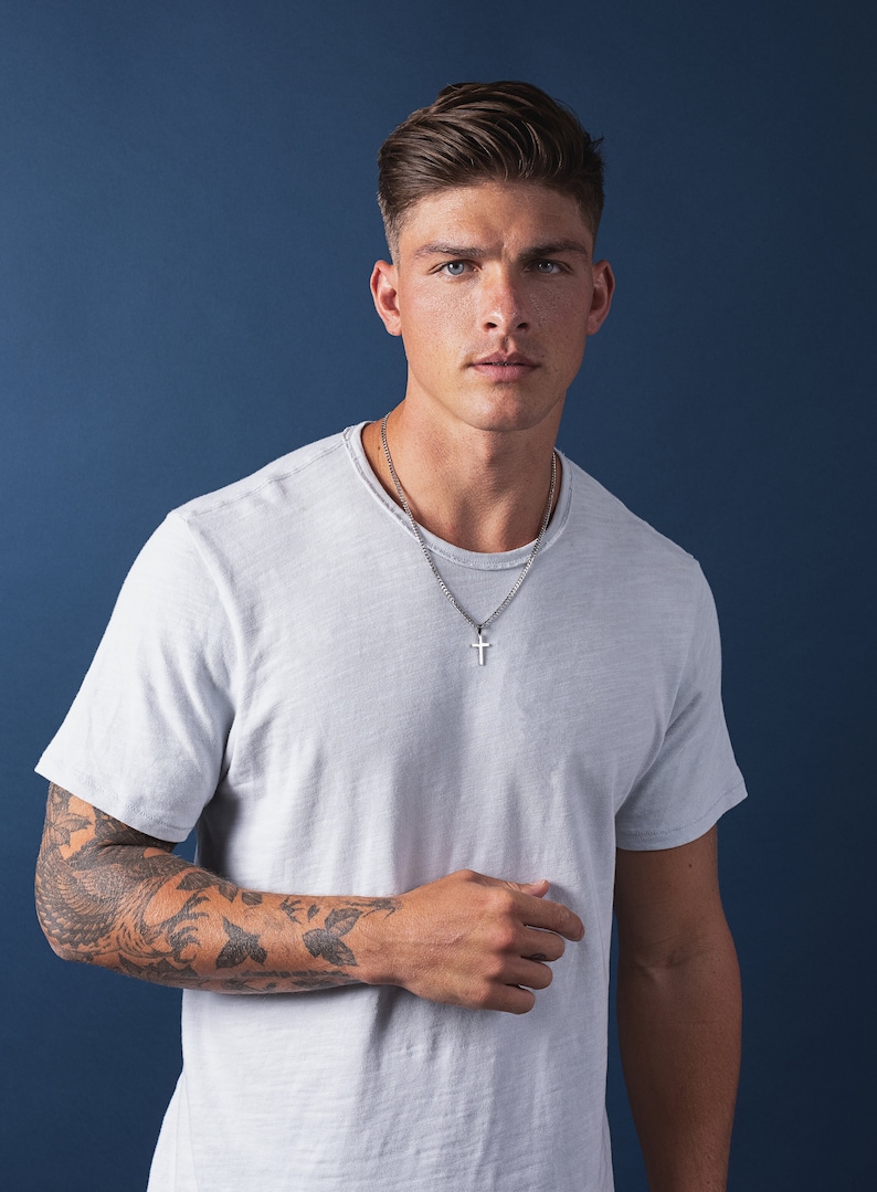 May include: A man wearing a white t-shirt with a silver chain necklace and a cross pendant. He has a tattoo on his left arm.