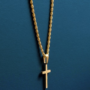 May include: Gold-colored necklace with a cross pendant. The chain has a rope design, and the cross is a simple, solid shape. The necklace hangs against a dark teal background.