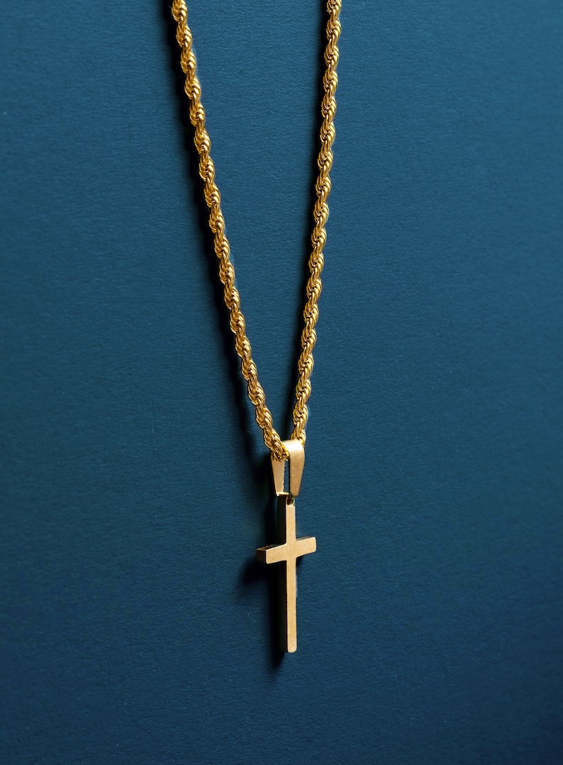 May include: Gold-colored necklace with a cross pendant. The chain has a rope design, and the cross is a simple, solid shape. The necklace hangs against a dark teal background.