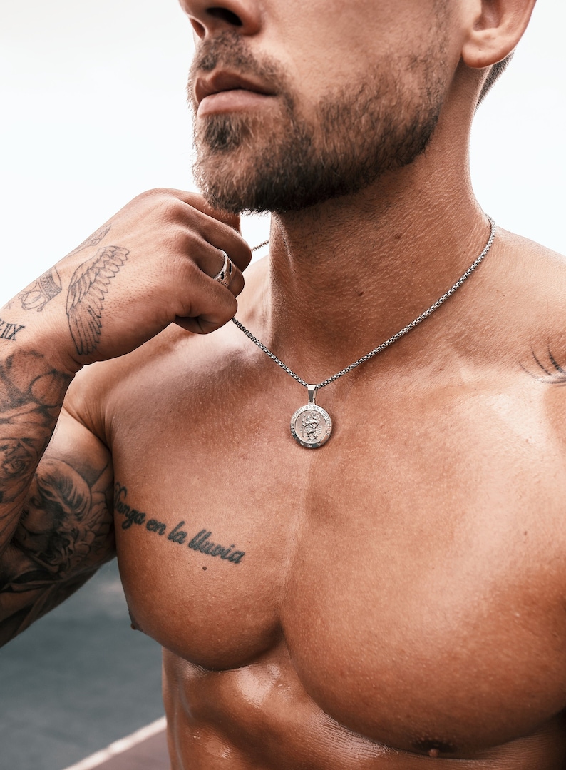 May include: A silver chain necklace with a round pendant featuring a detailed design. The pendant is hanging on a man's bare chest.