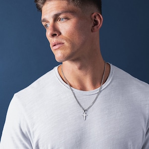 May include: A man wearing a white t-shirt with a silver chain necklace and a small silver cross pendant.