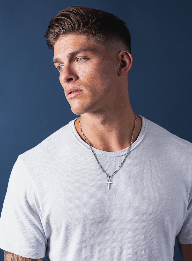 May include: A man wearing a white t-shirt with a silver chain necklace and a small silver cross pendant.