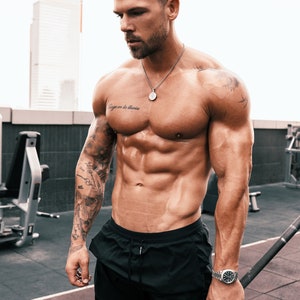 May include: A muscular man wearing black athletic shorts, a silver necklace with a pendant, and a silver watch. He has a tattoo on his left arm and a tattoo on his chest that says "Siempre en la memoria".