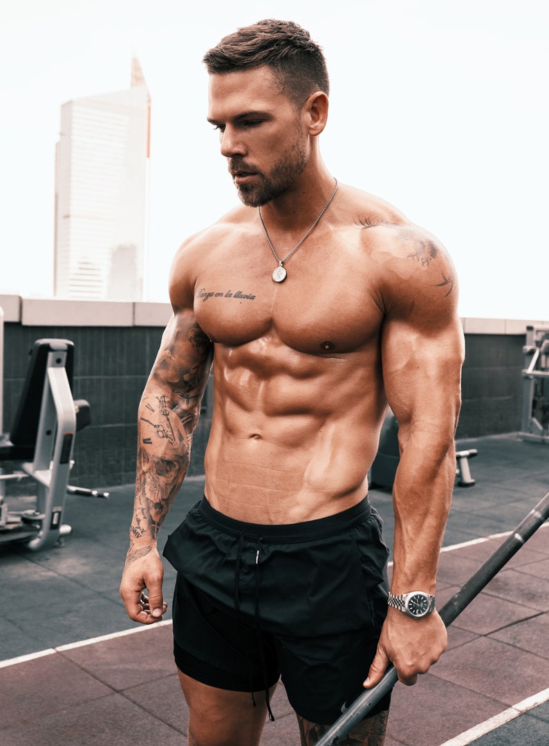 May include: A muscular man wearing black athletic shorts, a silver necklace with a pendant, and a silver watch. He has a tattoo on his left arm and a tattoo on his chest that says "Siempre en la memoria".