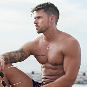 May include: A man wearing a black and gold patterned swim trunks with a gold chain necklace and a small gold cross pendant. The text "JOSS IS WEARING MEDIUM CROSS AT 20 INCHES" is printed on the bottom of the image.