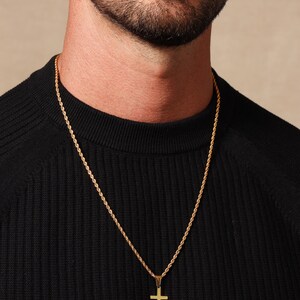 May include: Gold-colored cross pendant necklace. The pendant is a simple cross shape, suspended from a matching gold rope chain. The necklace is worn over a black sweater.