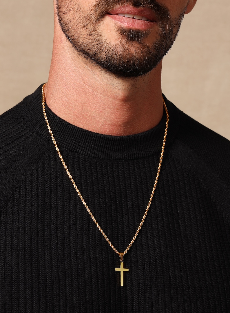 May include: Gold-colored cross pendant necklace. The pendant is a simple cross shape, suspended from a matching gold rope chain. The necklace is worn over a black sweater.