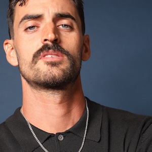 May include: A man with a moustache and beard is wearing a black polo shirt with a silver chain necklace.