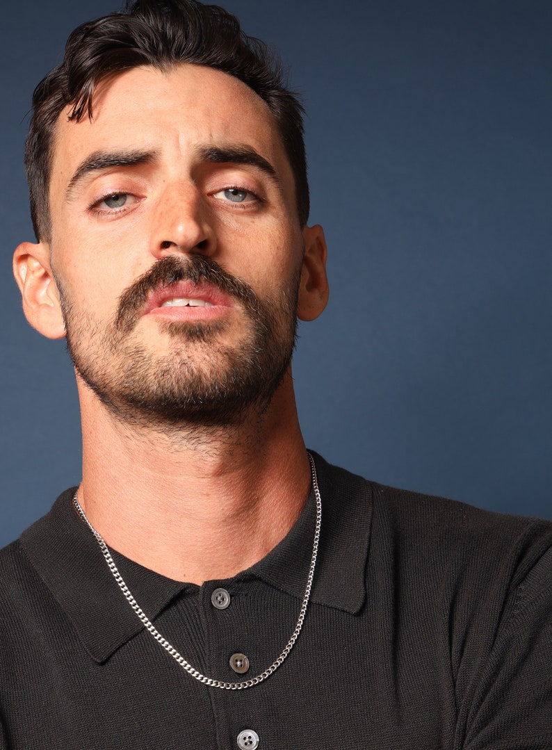 May include: A man with a moustache and beard is wearing a black polo shirt with a silver chain necklace.