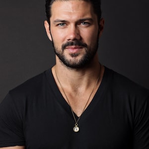 May include: A man with a beard and short brown hair is wearing a black v-neck t-shirt and a gold chain necklace with a compass pendant.