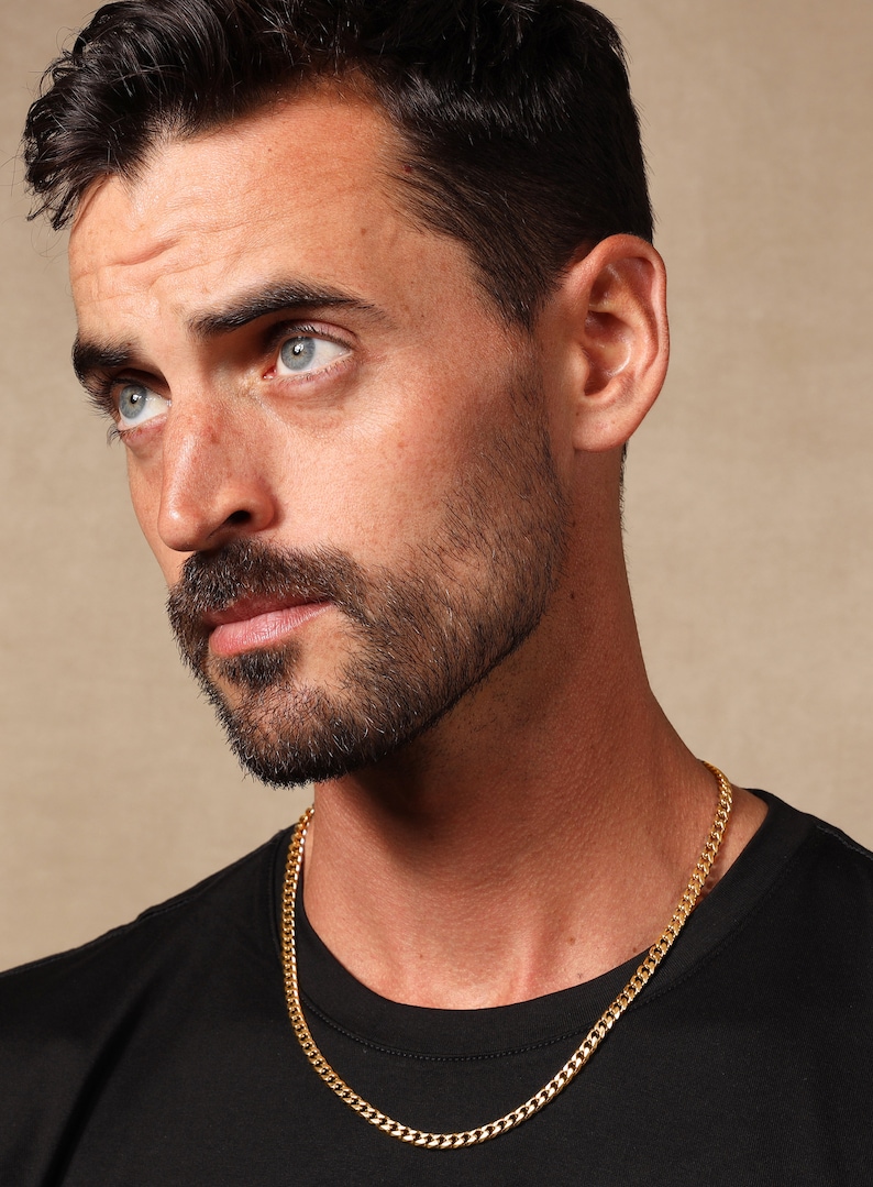May include: A gold chain necklace with a curb link style. The necklace is worn by a person with a beard and short dark hair.